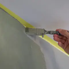 En hand målar en vägg inomhus med en pensel. Gul maskeringstejp skyddar taket.