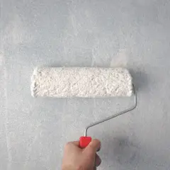 En hand håller en färgroller med vit färg, redo att måla på en ljusblå, texturerad tapet.