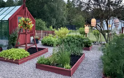 En lummig trädgård med röda pallkragar som inspirerar till odling. Grönsaksland och blommor samsas under hängande lyktor.