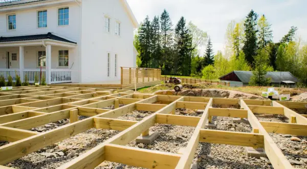 Grundläggande stomme för att bygga veranda framför ett vitt hus. Träbjälkar ligger på grus och stenar, redo för nästa steg.