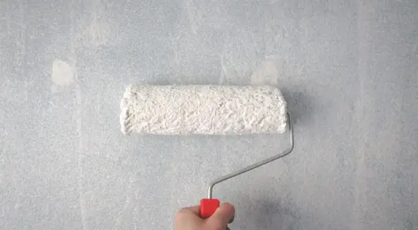 En hand håller en färgroller med vit färg, redo att måla på en ljusblå, texturerad tapet.