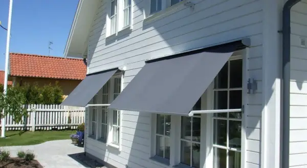 Grå markiser monterade på ett vitt trähus ger skugga åt fönstren.