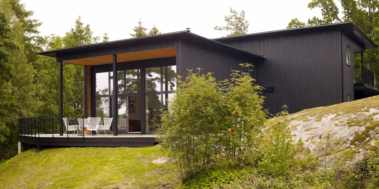 Modernt hus med mörkbrun fasad, stora fönster och en altan med vita stolar. Omgiven av grönskande träd och en mossbelagd sten.