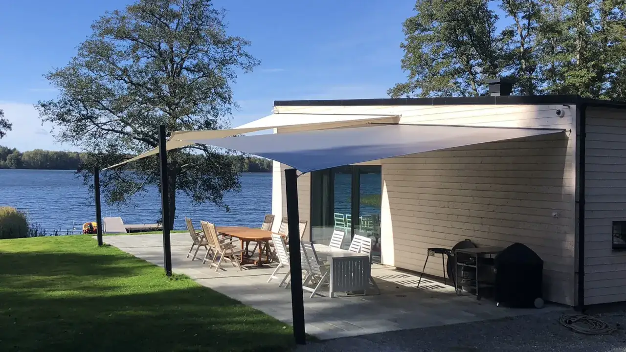 Solsegel ger skugga &ouml;ver uteplatsen vid sj&ouml;n. Tr&auml;dg&aring;rdsm&ouml;bler och grill st&aring;r redo f&ouml;r sommarens middagar.