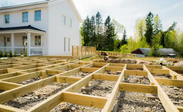 Grundläggande stomme för att bygga veranda framför ett vitt hus. Träbjälkar ligger på grus och stenar, redo för nästa steg.