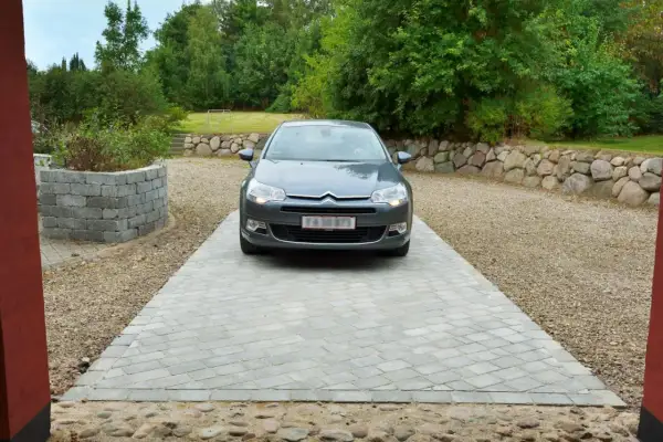 Grå bil parkerad på en vacker betong uppfart. Omgiven av grönskande träd och en stenmur.