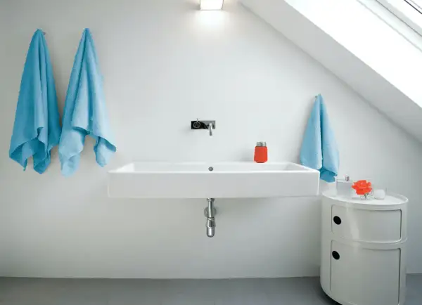 Modernt badrum med ett stilrent handfat. Två ljusblåa handdukar hänger på vänster sida, en på höger. En orange behållare och en blomma står på ett litet vitt skåp.
